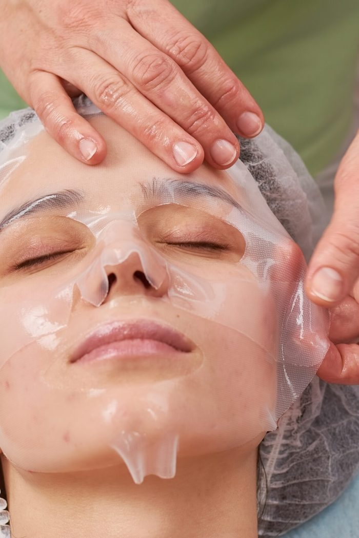 Hands applying collagen facial mask. Face of young woman, cosmetology. Getting rid of acne.